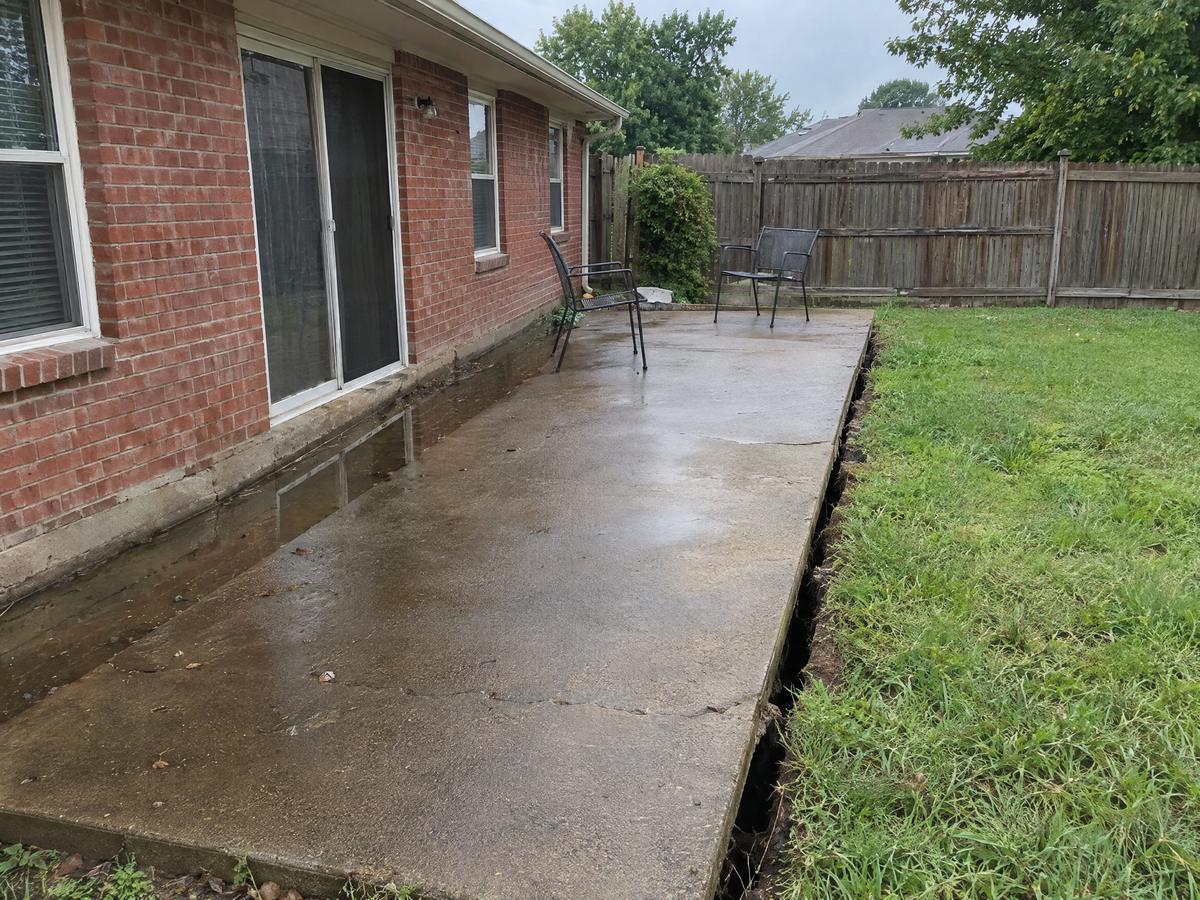 A backyard concrete patio behind a brick ranch house that has tipped toward the foundation, with rainwater pooled along the house wall
