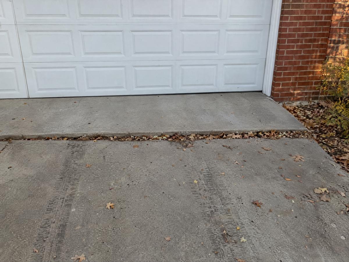 A driveway in front of a white garage door where the outer panel has dropped below the apron, leaving a leaf-filled step and tire tracks on the low slab