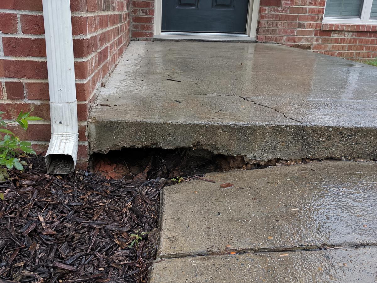 A concrete front stoop with soil washed out from under its edge beside a downspout, leaving a dark hollow under the slab